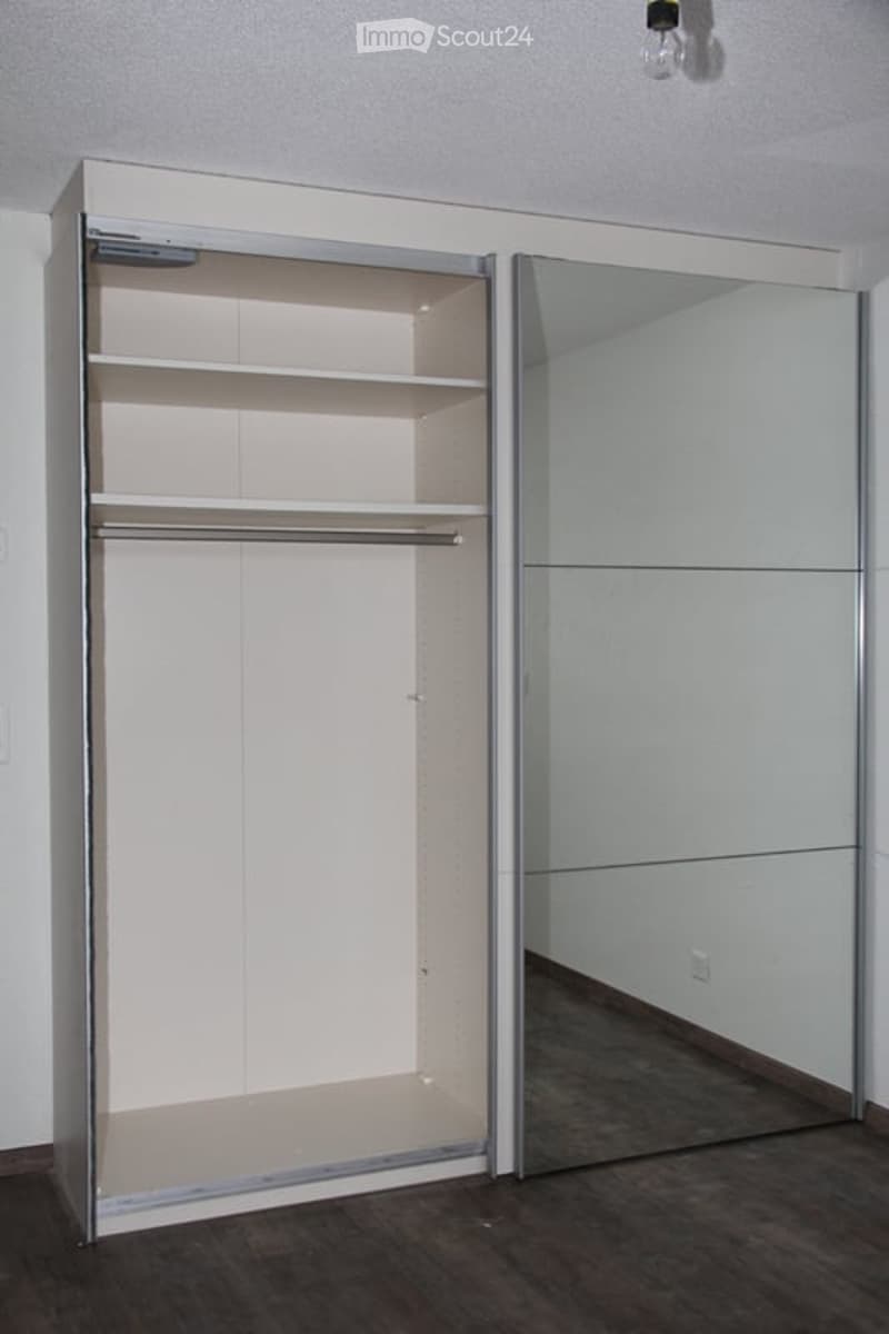 Empty room with a large, modern walk-in closet featuring mirrored sliding doors, multiple shelves, and a hanging rod. The room has a dark wooden floor and white walls.