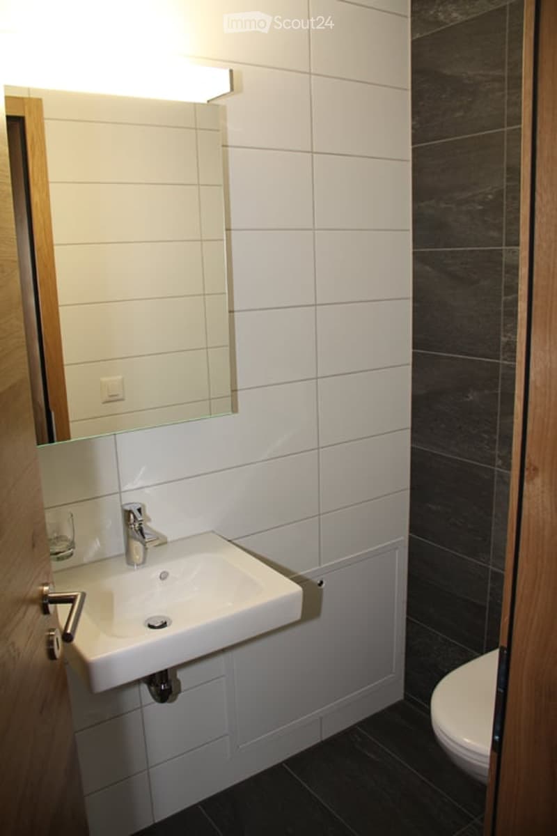 Modern bathroom with white tiles, a white sink, a mirror, and a toilet. The walls are tiled in white and dark grey, and the floor is dark grey.