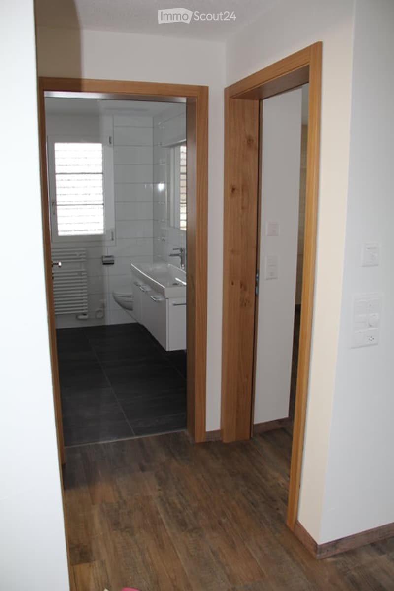 A hallway with wooden flooring and white walls, leading to a bathroom with white tiles, a sink, and a toilet.