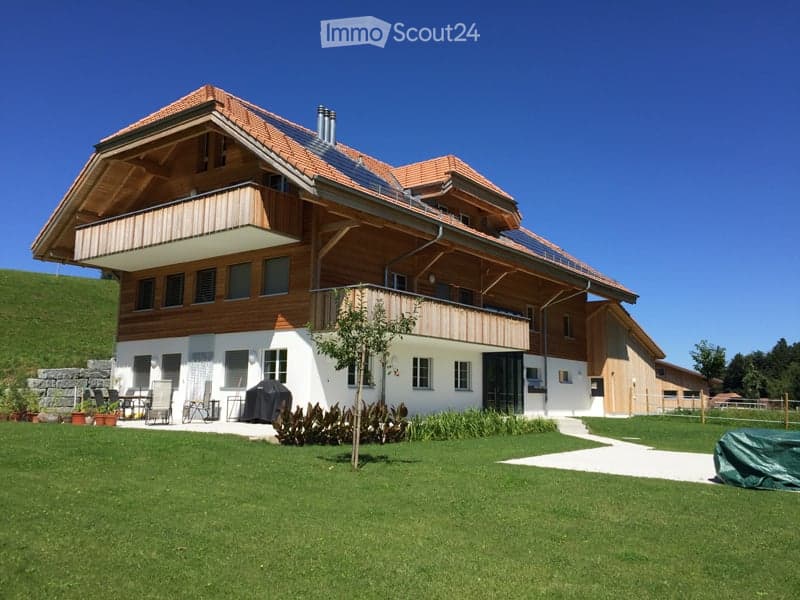 Modern two-story house with wooden balconies, solar panels on the roof, and a well-maintained lawn in front.
