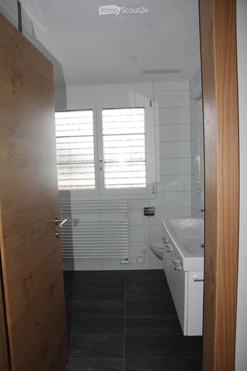 Modern bathroom with dark tiled floor, white walls, a sink, toilet, radiator, and a large window with blinds.