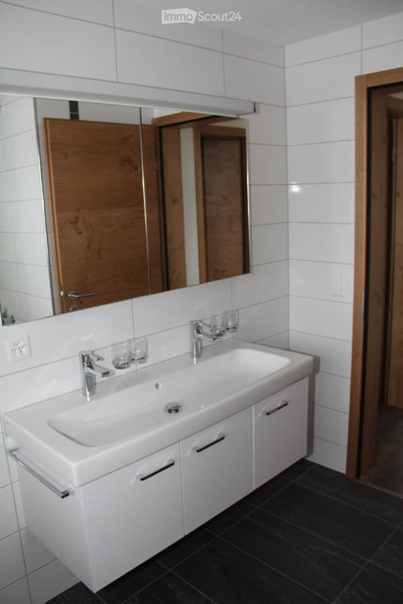 Modern bathroom with white tiles, a large sink with a mirror above it, and a wooden door in the background.