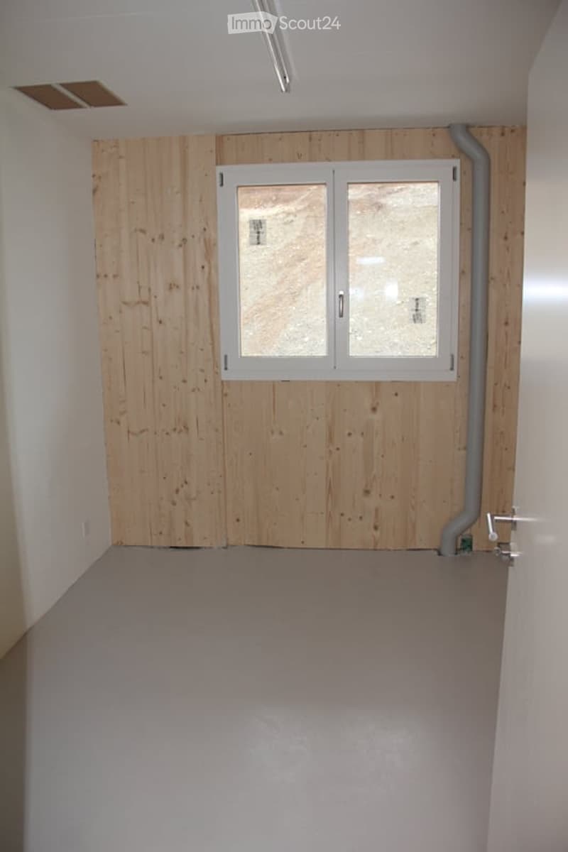 Empty room with wooden panel wall, white walls, grey floor, and a window with white frame.