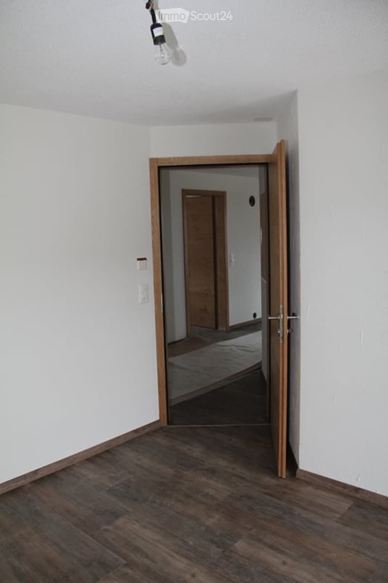 Empty room with wooden flooring, white walls, and a partially open wooden door leading to another room.