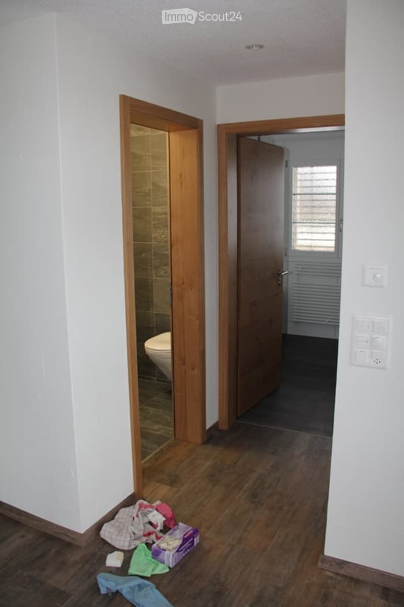 Empty hallway with wooden flooring, white walls, and open doors leading to a bathroom and another room. Some scattered items are on the floor near the entrance.