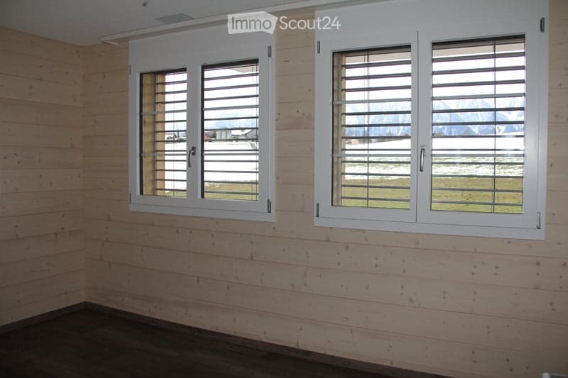 Empty room with wooden walls and flooring, featuring two large windows with white shutters overlooking a snowy landscape.