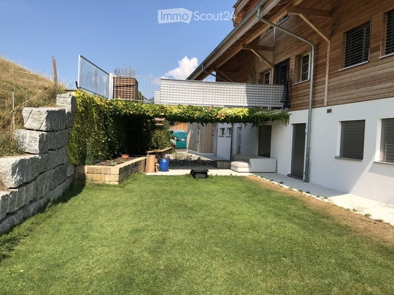Well-maintained garden with a neatly trimmed lawn, stone retaining wall, and a covered seating area with a glass railing. The area is bordered by a wooden house with large windows and a balcony.