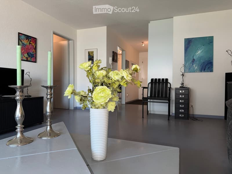 Modern living room with a grey table, white vase with yellow flowers, silver candle holders, black chair, and framed artwork on the walls.