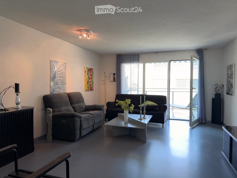 Modern living room with two sofas, a coffee table, a chair, and decorative items including candles and a vase with flowers. Large windows and a sliding glass door lead to a balcony.