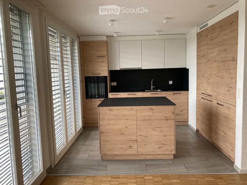 Modern kitchen with light wood cabinets, a central island, and a black countertop. Large windows with blinds provide natural light.