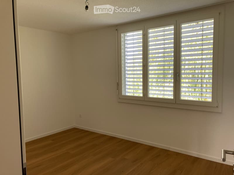 Empty room with light wooden flooring, white walls, and large windows with blinds. No furniture or equipment present.