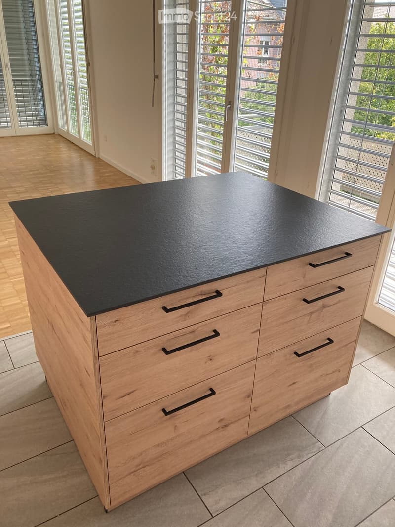 Modern kitchen island with light wood drawers and a black countertop, situated near large windows with blinds and a view of the outdoor area.
