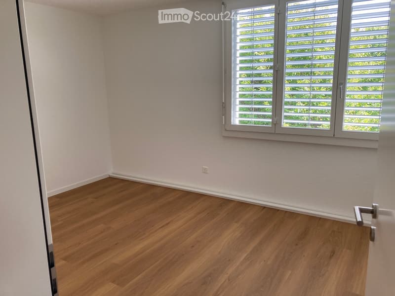 Empty room with light-colored walls, wooden flooring, and a large window with blinds.