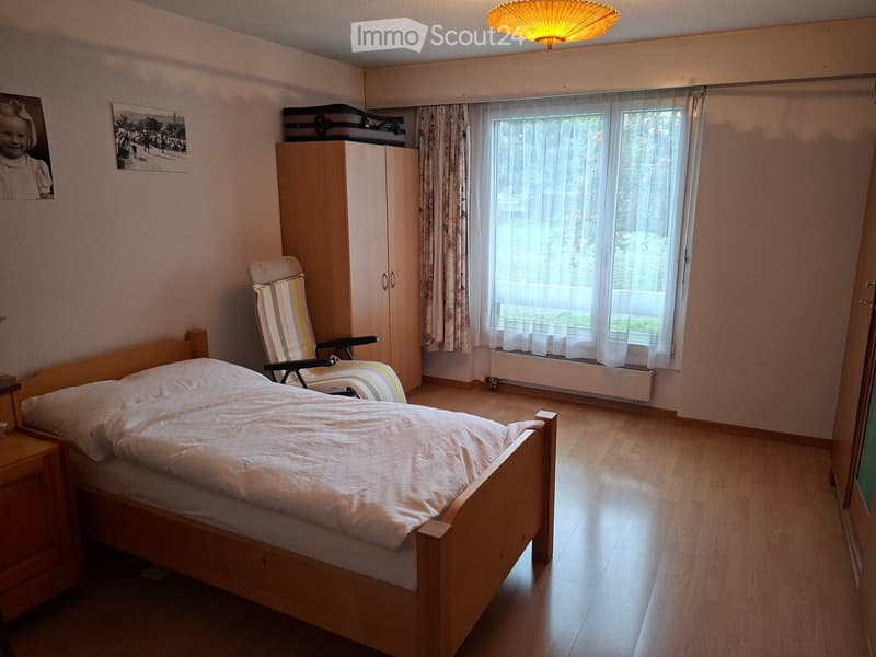Cozy bedroom with wooden bed, nightstand, wardrobe, and a comfortable chair near the window.