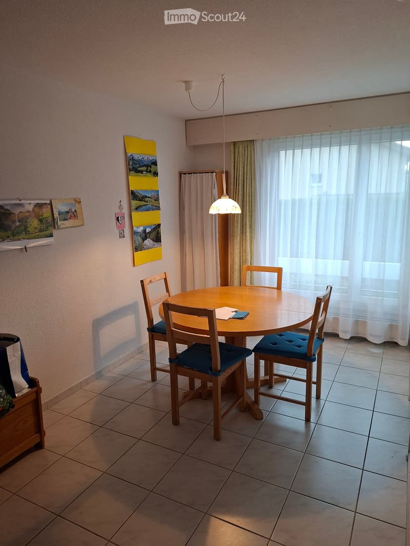 A small kitchen with a round wooden dining table surrounded by four wooden chairs with blue cushions, a pendant light hanging above, and a window with white curtains. The walls are decorated with picture frames and a calendar.