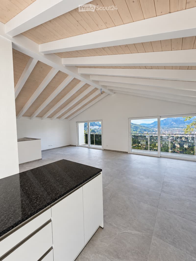 Open-plan space with white walls, light wooden ceiling beams, large windows offering a scenic view, and a modern kitchen island with a black countertop.