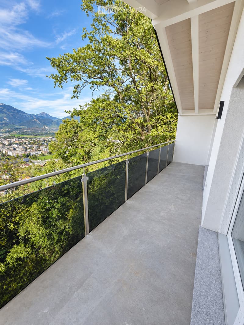 Modern balcony with glass railing, overlooking a lush green landscape and distant mountains.