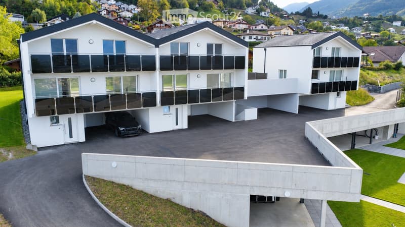 Modern two-story white house with large windows and balconies, situated in a scenic valley with surrounding houses and mountains.