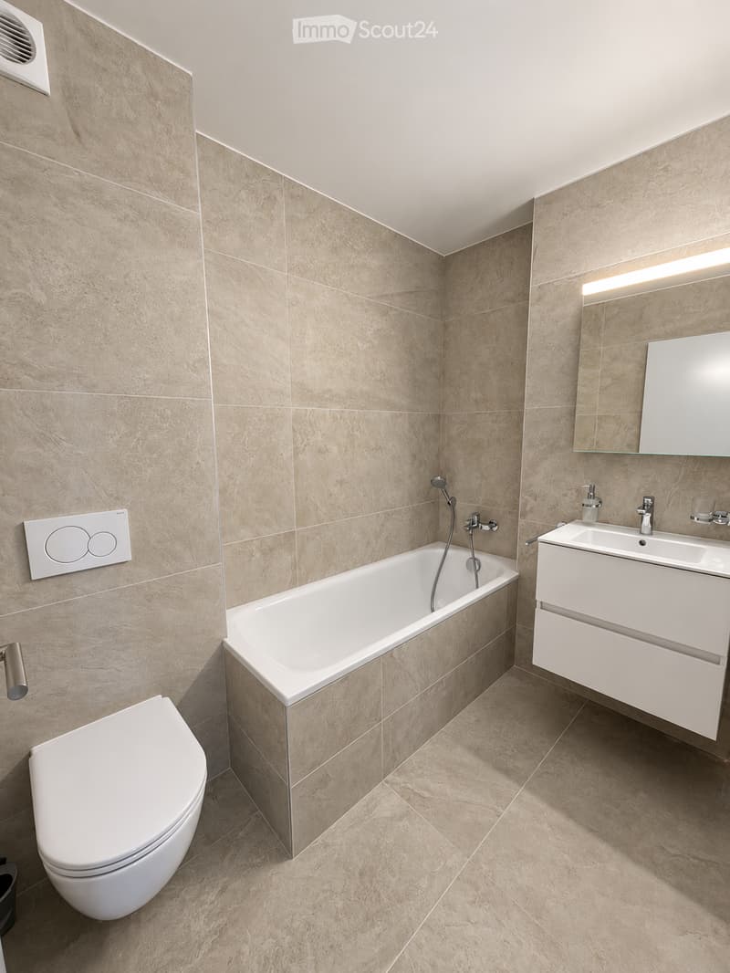 Modern bathroom with beige tiled walls and floor, featuring a bathtub, toilet, sink, and a large mirror above the sink.