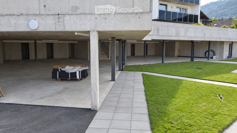 Modern concrete building with a covered parking area, grassy lawn, and a raised ground floor section.