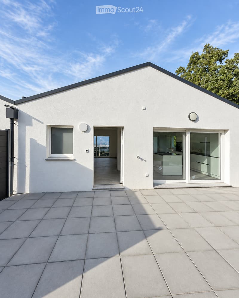 Modern white house with a flat roof, large windows, and a spacious patio area. The house features a sleek design with minimalistic architecture and a clean, contemporary aesthetic.