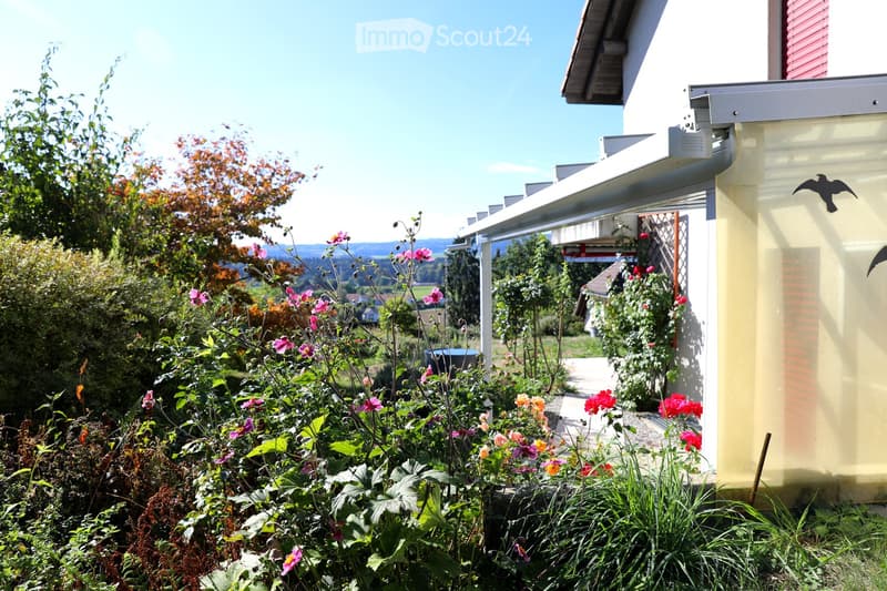 A well-maintained garden with colorful flowers, shrubs, and a pathway leading to a house with a covered entrance. The background shows a scenic view of hills and trees.
