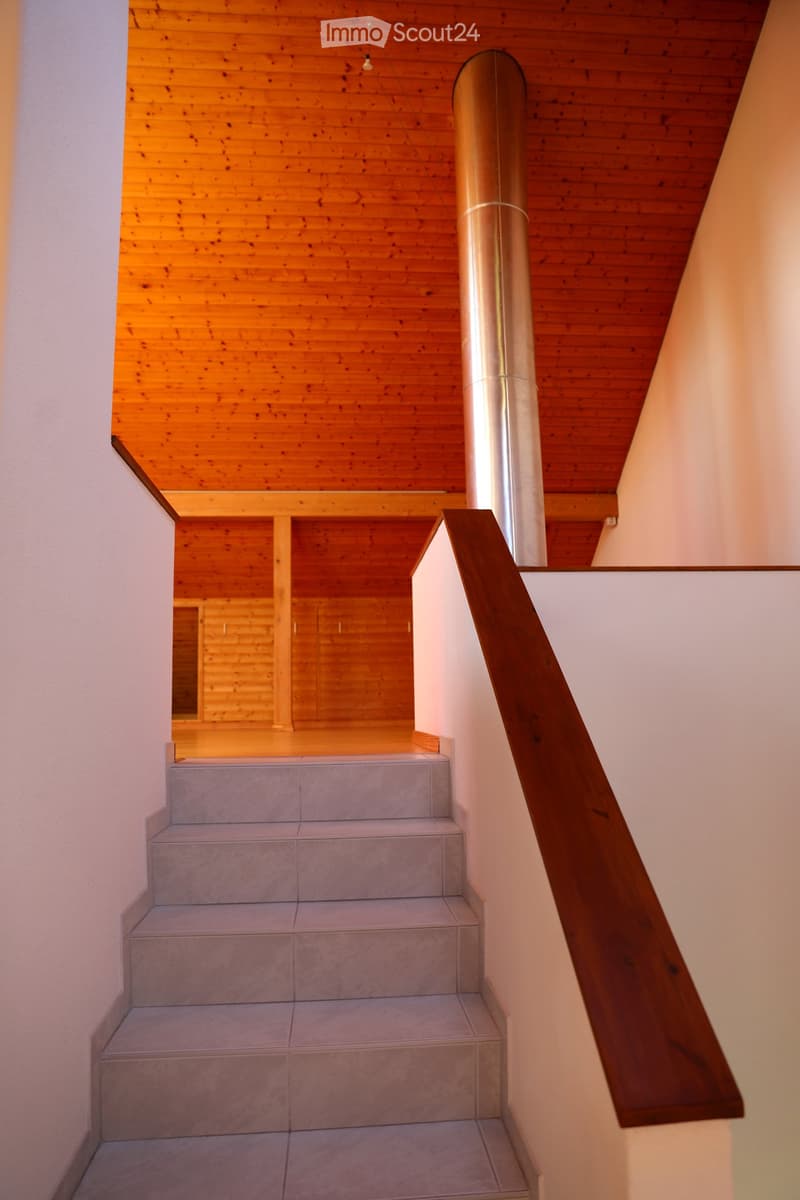 Wooden staircase with beige tiled steps and a wooden handrail, leading to a wooden ceiling area with warm lighting.