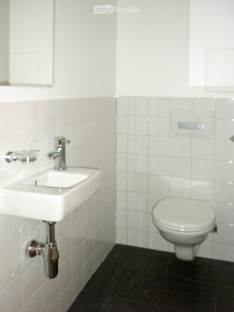 Modern bathroom with white tiled walls, a white sink with a chrome faucet, and a white toilet. Dark tiled floor.
