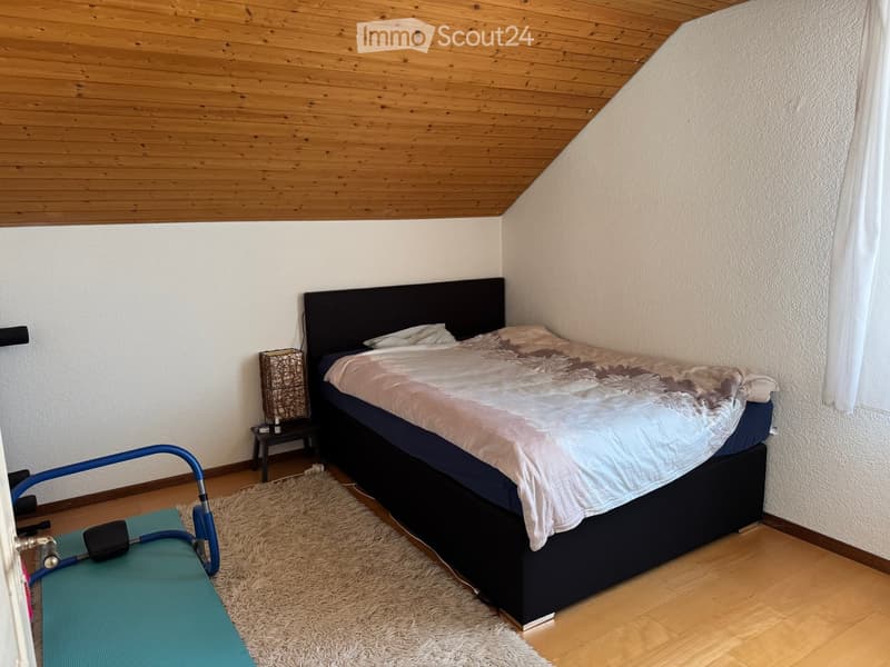 Cozy bedroom with a wooden slanted ceiling, a neatly made bed with a dark headboard, a nightstand with a lamp, and a shaggy rug on the floor.