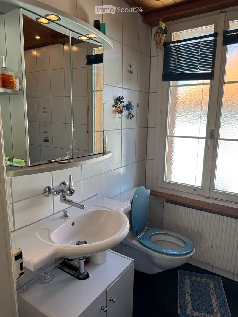 Modern bathroom with a white sink, chrome faucet, toilet with a blue seat, large mirror with built-in lighting, and a window with blinds.