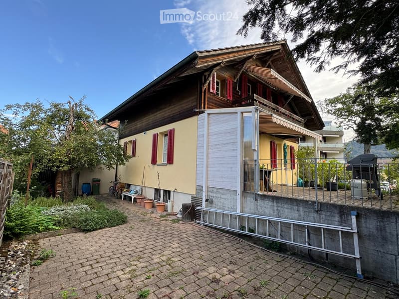 Two-story house with a wooden upper level, yellow lower level, red shutters, and a covered balcony area. The property includes a paved patio, garden, and outdoor furniture.