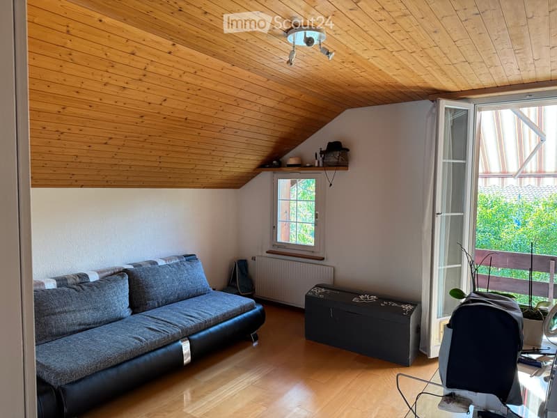 A cozy living room with wooden floors, a slanted wooden ceiling, large windows, a grey leather sofa, a small table, a chair, and a plant near the window.
