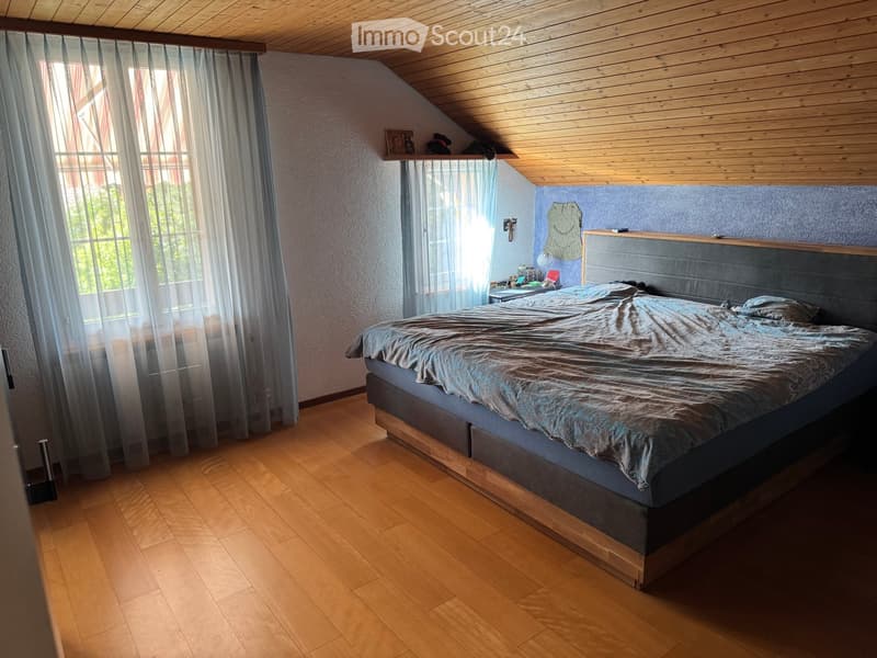 Cozy attic bedroom with wooden slanted ceiling, hardwood floor, large window with sheer curtains, bed with grey headboard, nightstand, and small shelf on the wall.