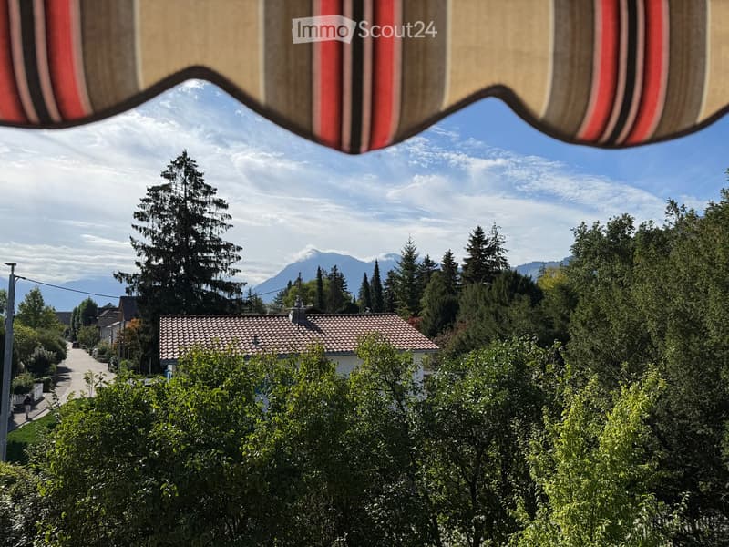 View from a balcony or terrace showing a suburban neighborhood with houses, trees, and distant mountains under a clear sky.