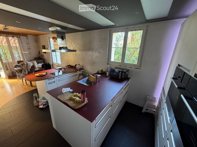 Modern kitchen with white cabinets, red countertop, and stainless steel appliances. Features a central island with sink, microwave, and various kitchen items.