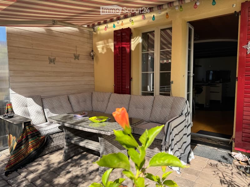 Outdoor patio with a sofa, table, and bench, decorated with a colorful blanket and a flower in the foreground.