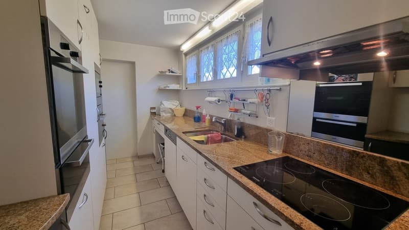 Modern kitchen with white cabinets, granite countertops, stainless steel appliances, and a window with blinds. Features an induction stove, refrigerator, and sink with cleaning supplies on the counter.