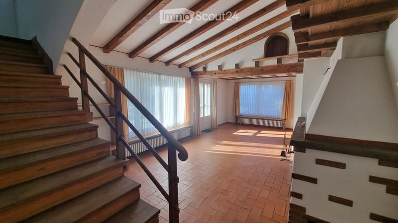 Spacious hallway with wooden staircase, exposed wooden beams, large windows with curtains, and tiled floor.