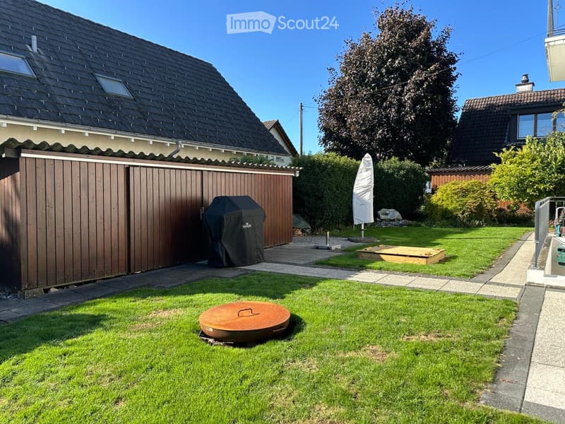 Garden with brown wooden fence, barbecue, round wooden bench, closed white umbrella, grassy area, metal grate, and another house in the background.