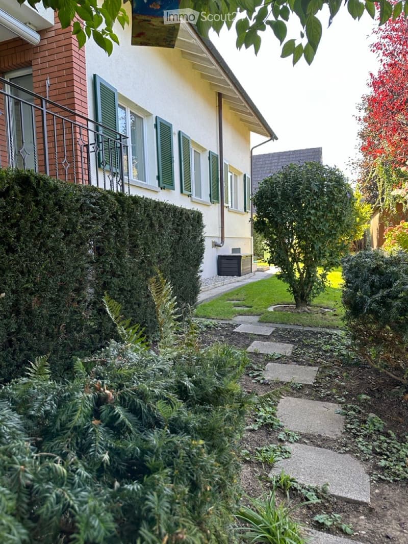 House exterior with brick wall, green window shutters, balcony, well-maintained garden, small pathway