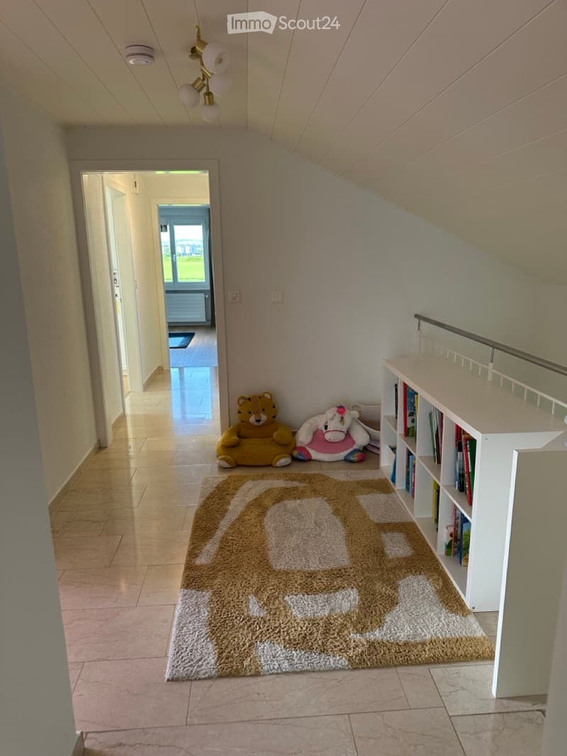 Tiled floor, white walls, decorative rug, two stuffed toys, a white bookcase, an open door, a ceiling with a light fixture and a smoke detector, a small rug on the floor.