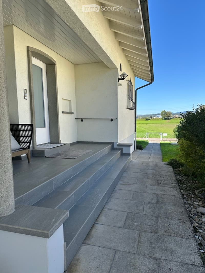 Modern house entrance with stone steps, tiled pathway, grey stone columns, green field view.
