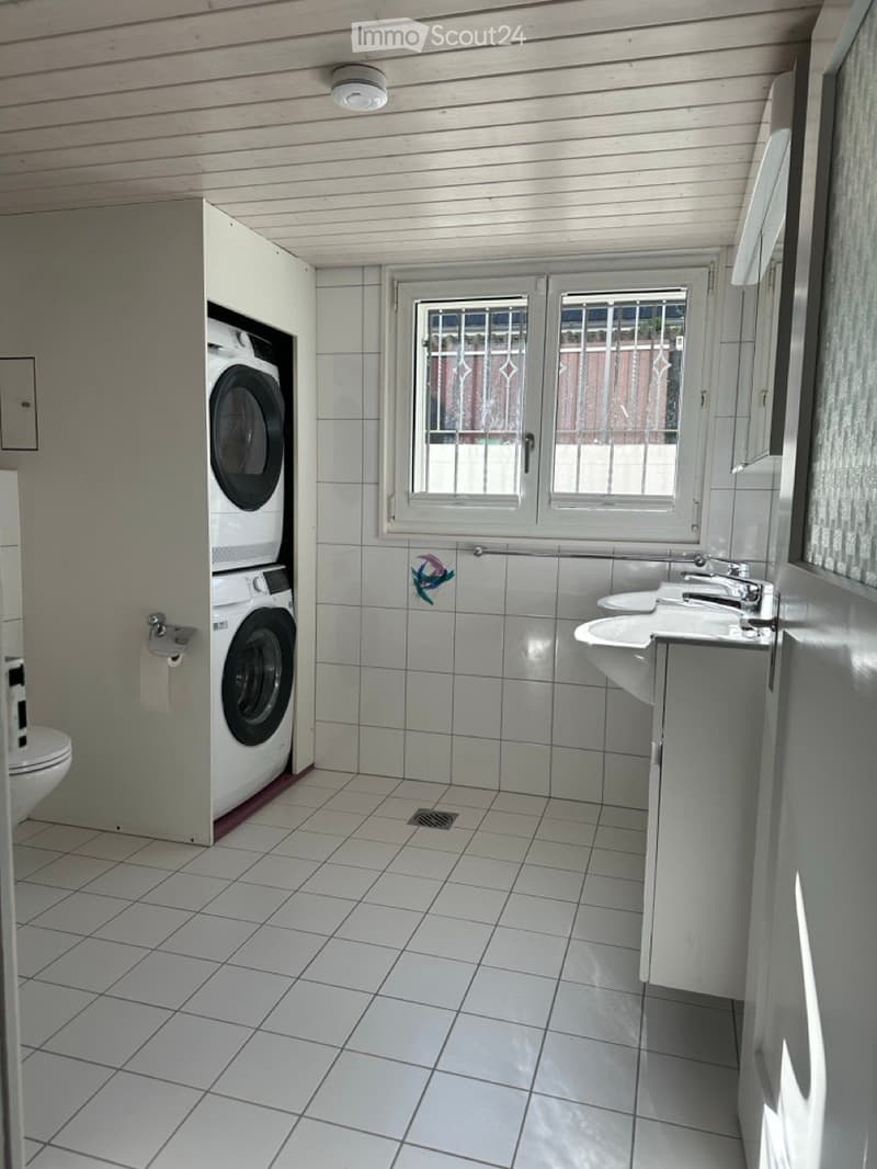 Bathroom with white tiled floor, two washing machines, two sinks, white toilet, white cabinet, window