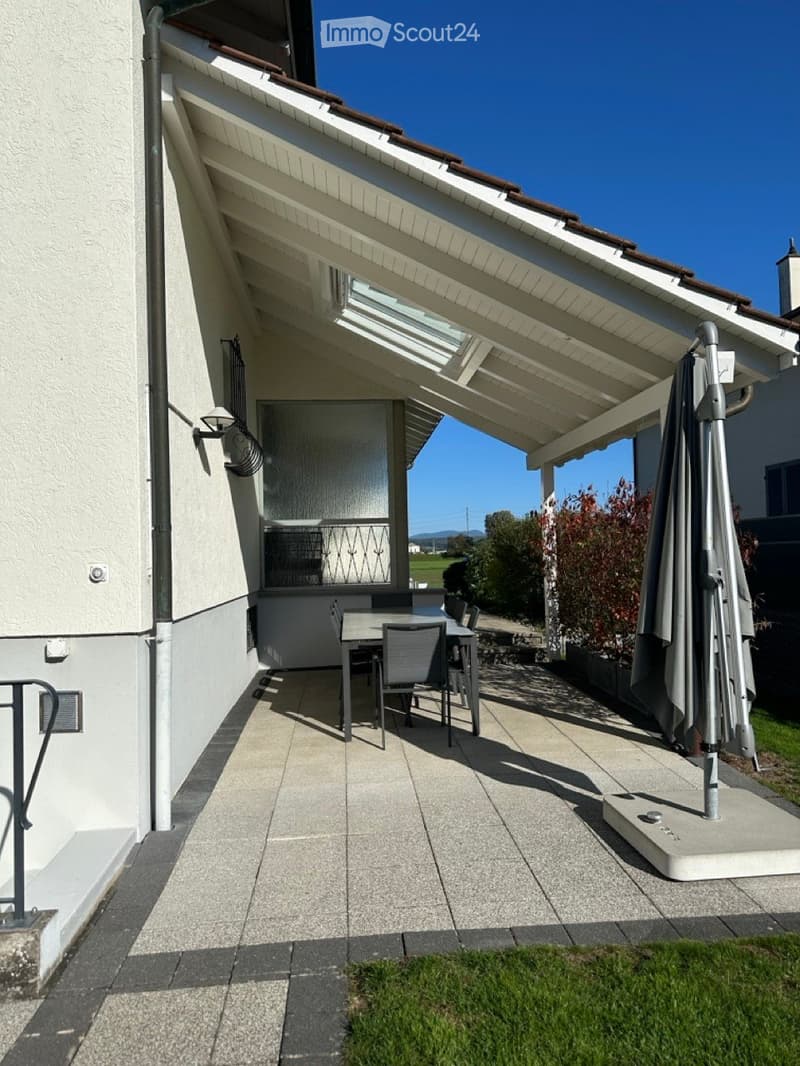 Outdoor patio covered by a pergola, glass window, glass doors, wooden table and chairs, parasol, light fixtures, plants and greenery, tiled and gravel floor