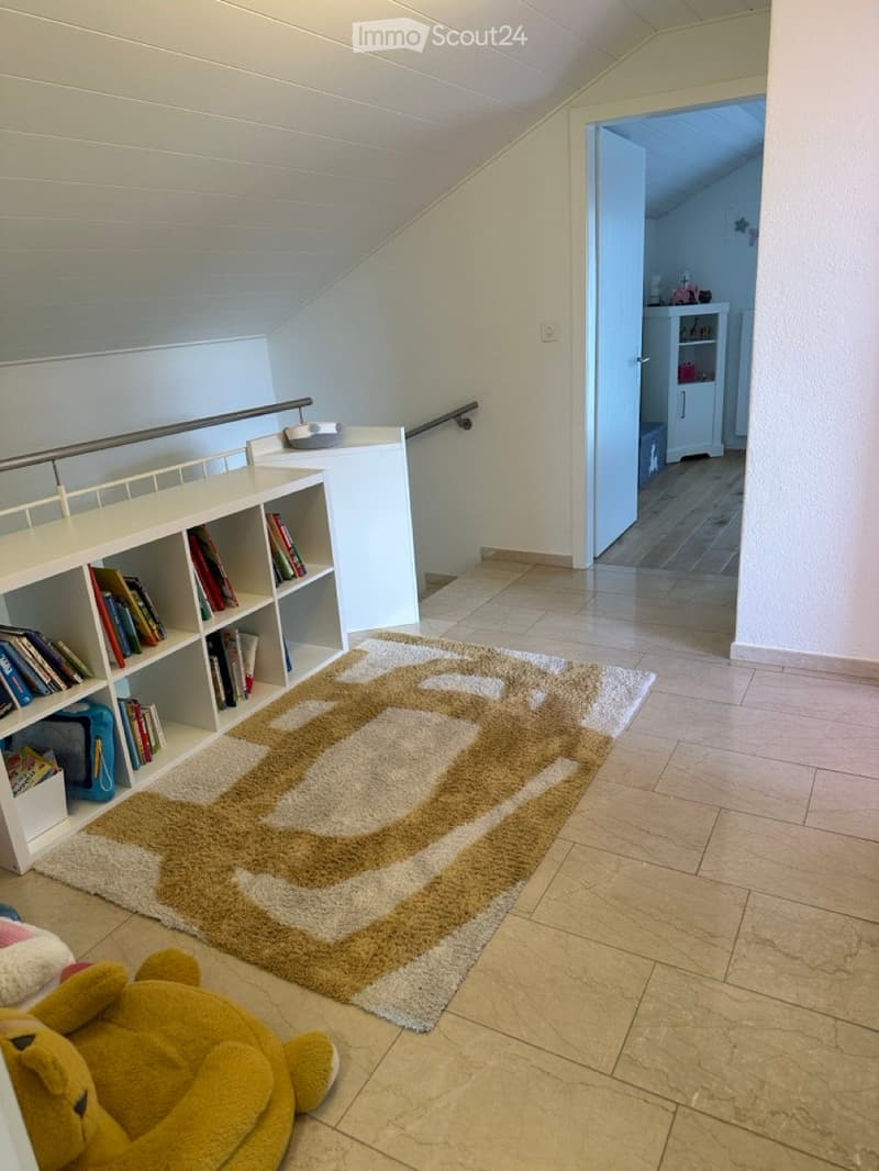 Attic room with beige tiled floor, white bookshelves, beige rug, white walls, brown teddy bear, shelves filled with books.