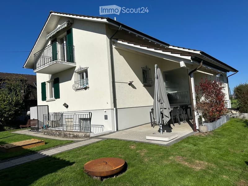 Two story house, white wall, brown roof, patio, green shutters, wooden balcony, green grass, iron fence, table and chairs on patio