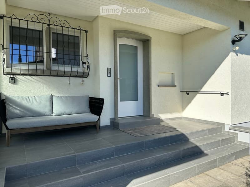 Raised ground floor, bench with cushions, entry door with a mat, wrought iron balcony railing, mounted lamp, white textured walls