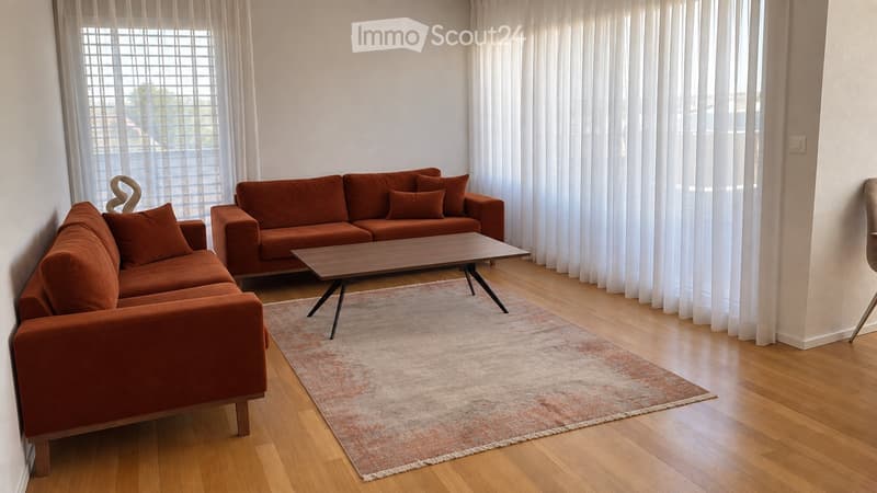 Modern living room with two orange sofas, a wooden coffee table, and a patterned rug on a wooden floor. Large windows with white curtains provide natural light.