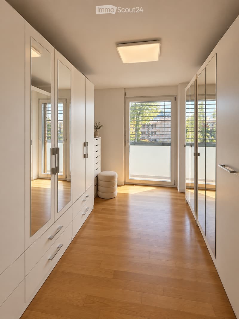 Modern hallway with light wooden flooring, white walls, large mirrored cabinets, a small round stool, and a vase with greenery near the entrance.