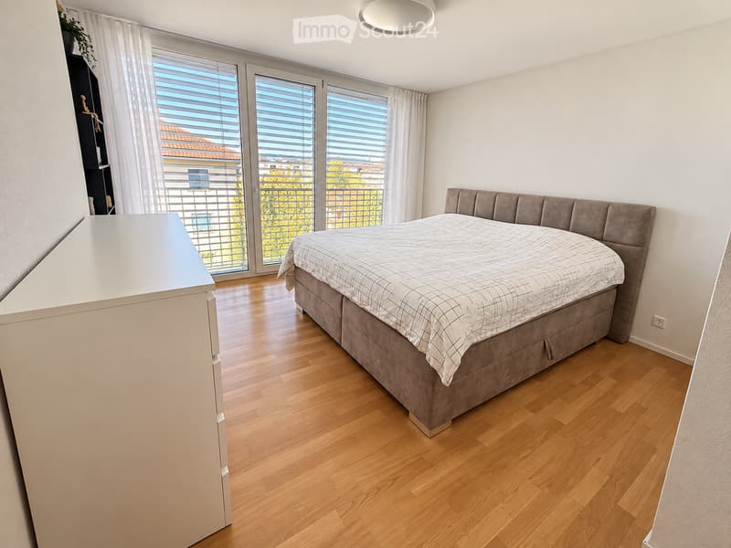 Modern bedroom with wooden flooring, large windows with blinds, grey upholstered bed, nightstand, and shelves with decorative items.